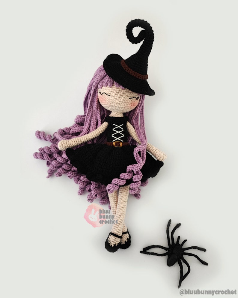 May include: A crocheted witch doll with purple hair, a black dress, and a black witch hat. The doll is lying on its back with its legs crossed. A black crocheted spider is on the white surface below the doll. The doll's eyes are closed and it has a sweet expression. The doll is wearing a black belt with a buckle. The doll's hair is in a curly style. The doll is made by @bluubunnycrochet.