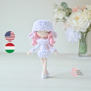 May include: White crocheted doll with pink hair, wearing a white dress and white shoes. The doll is standing on a white surface. The background is a white wall with a vase of white flowers.
