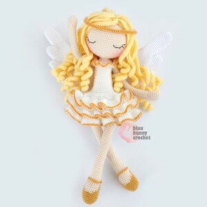 May include: A crocheted angel doll with blonde curly hair, a white dress with gold trim, and white wings. The doll is wearing gold shoes and has a gold halo. The doll is lying on its back with its arms raised above its head. The doll is made by "bluu bunny crochet".