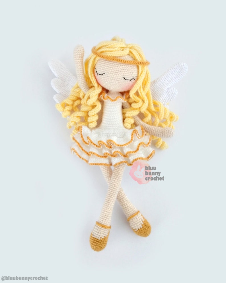 May include: A crocheted angel doll with blonde curly hair, a white dress with gold trim, and white wings. The doll is wearing gold shoes and has a gold halo. The doll is lying on its back with its arms raised above its head. The doll is made by "bluu bunny crochet".