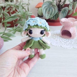 Puede incluir: Mu&ntilde;eca de ganchillo hecha a mano con una corona de flores azul claro y un vestido verde. La mu&ntilde;eca tiene grandes ojos negros y orejas peque&ntilde;as. El texto "bluu bunny croche" es visible en una etiqueta.