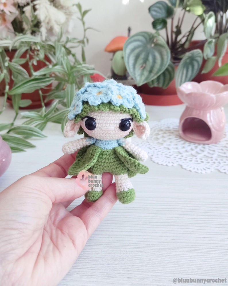 Puede incluir: Mu&ntilde;eca de ganchillo hecha a mano con una corona de flores azul claro y un vestido verde. La mu&ntilde;eca tiene grandes ojos negros y orejas peque&ntilde;as. El texto "bluu bunny croche" es visible en una etiqueta.