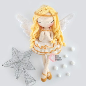 May include: A crocheted angel doll with blonde hair, white wings, and a white dress with a gold trim. The doll is holding its hands together in prayer. The doll is sitting on a white surface with a silver star and white pom-poms.