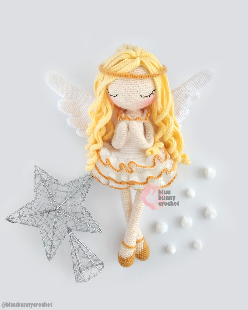 May include: A crocheted angel doll with blonde hair, white wings, and a white dress with a gold trim. The doll is holding its hands together in prayer. The doll is sitting on a white surface with a silver star and white pom-poms.