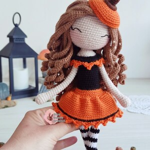 May include: Crocheted doll dressed in an orange and black Halloween costume with a pumpkin hat. The doll has brown curly hair and is holding a small crocheted pumpkin.