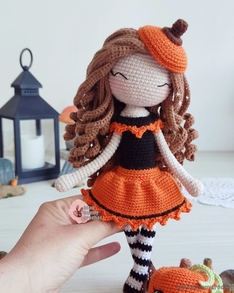 May include: Crocheted doll dressed in an orange and black Halloween costume with a pumpkin hat. The doll has brown curly hair and is holding a small crocheted pumpkin.