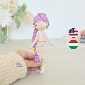 May include: Handmade crocheted doll with a purple braided hairstyle, gold detailing, and a white outfit. The doll has closed eyes and a delicate design. The doll is held by a person. The doll is handmade.