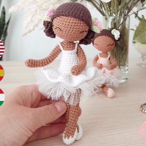 May include: Two crocheted ballerina dolls, one wearing a white tutu and the other wearing a pink tutu. Both dolls have brown skin and black hair.