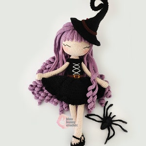 May include: A crocheted witch doll with long purple hair, wearing a black dress and a black witch hat with a curled brim. The doll is holding a black spider.