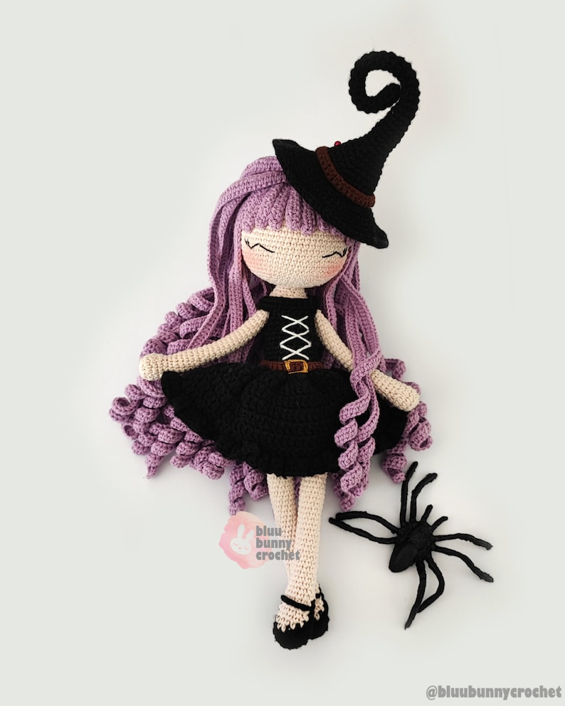 May include: A crocheted witch doll with long purple hair, wearing a black dress and a black witch hat with a curled brim. The doll is holding a black spider.