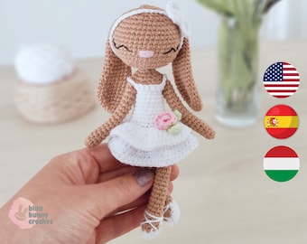 May include: A crocheted bunny doll wearing a white dress with a pink flower. The bunny has brown fur and a white headband. The doll is holding a pink flower in its hand.