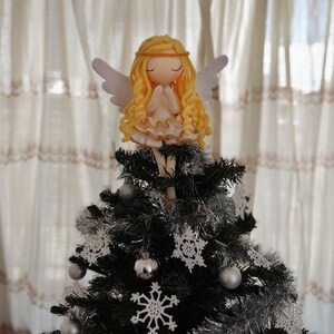May include: A crocheted angel doll with blonde hair and white wings sits atop a Christmas tree decorated with silver tinsel and white crocheted snowflakes.