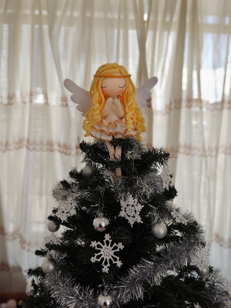 May include: A crocheted angel doll with blonde hair and white wings sits atop a Christmas tree decorated with silver tinsel and white crocheted snowflakes.