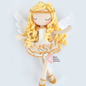 May include: A crocheted angel doll with blonde hair, white wings, and a white dress with gold trim. The doll is lying on its back with its eyes closed. The doll is wearing gold shoes.