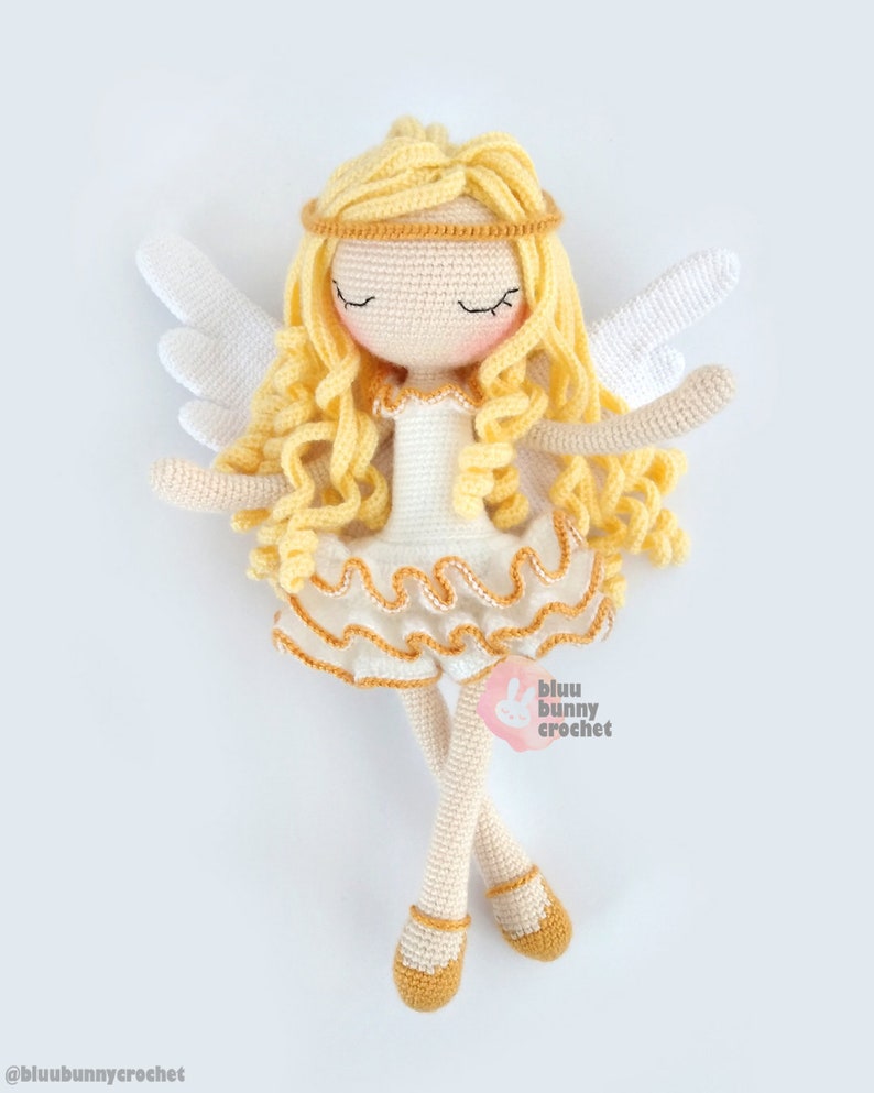 May include: A crocheted angel doll with blonde hair, white wings, and a white dress with gold trim. The doll is lying on its back with its eyes closed. The doll is wearing gold shoes.