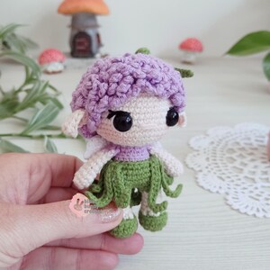 Puede incluir: Una mu&ntilde;eca de hada de ganchillo hecha a mano con una gorra morada, una cara blanca y una falda verde. La mu&ntilde;eca tiene ojos negros y peque&ntilde;as alas blancas. El hada sostiene una peque&ntilde;a hoja verde. El fondo incluye setas en miniatura y una casita.