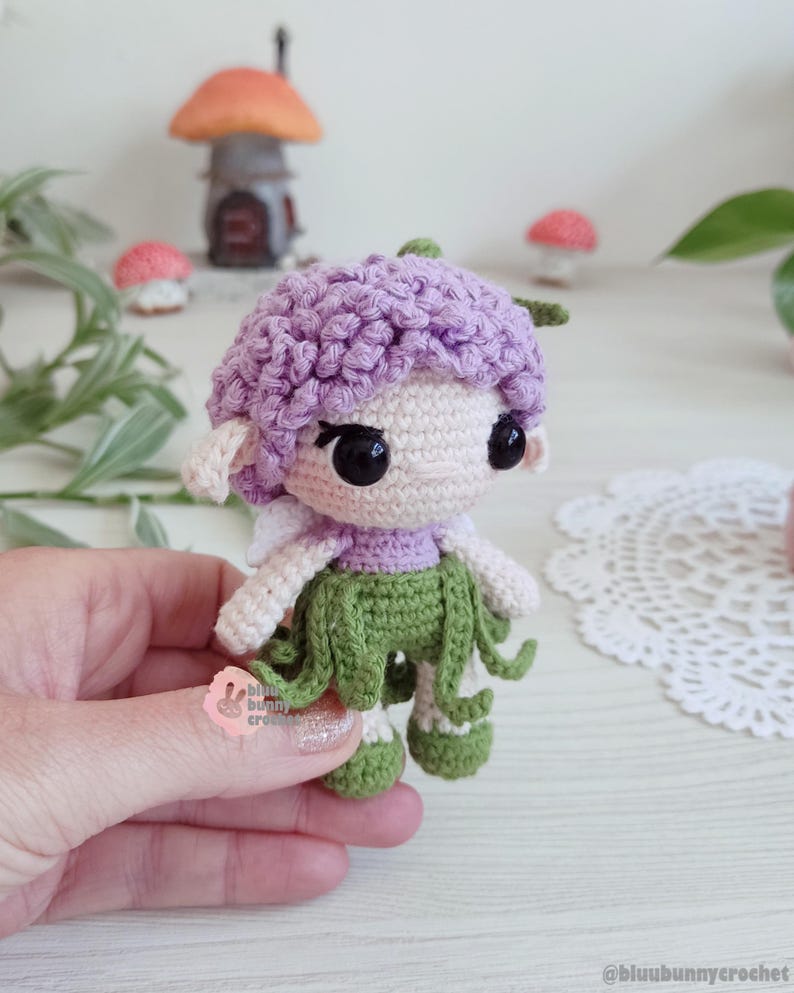 Puede incluir: Una mu&ntilde;eca de hada de ganchillo hecha a mano con una gorra morada, una cara blanca y una falda verde. La mu&ntilde;eca tiene ojos negros y peque&ntilde;as alas blancas. El hada sostiene una peque&ntilde;a hoja verde. El fondo incluye setas en miniatura y una casita.
