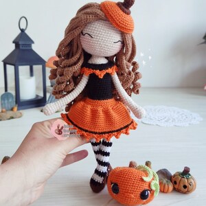 May include: A crocheted doll dressed in an orange and black Halloween costume. The doll has brown curly hair and is wearing a pumpkin hat. The doll is holding a small crocheted pumpkin.