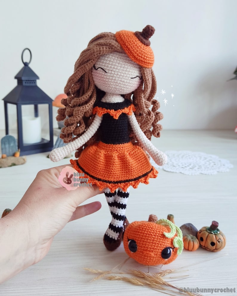 May include: A crocheted doll dressed in an orange and black Halloween costume. The doll has brown curly hair and is wearing a pumpkin hat. The doll is holding a small crocheted pumpkin.