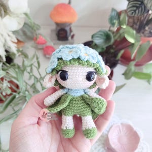 Puede incluir: Mu&ntilde;eca de ganchillo hecha a mano con una corona de flores y un vestido verde. La mu&ntilde;eca tiene ojos negros grandes, orejas peque&ntilde;as y un cuello azul claro. La mu&ntilde;eca se sostiene en una mano, con un plato rosa en forma de flor en el fondo.