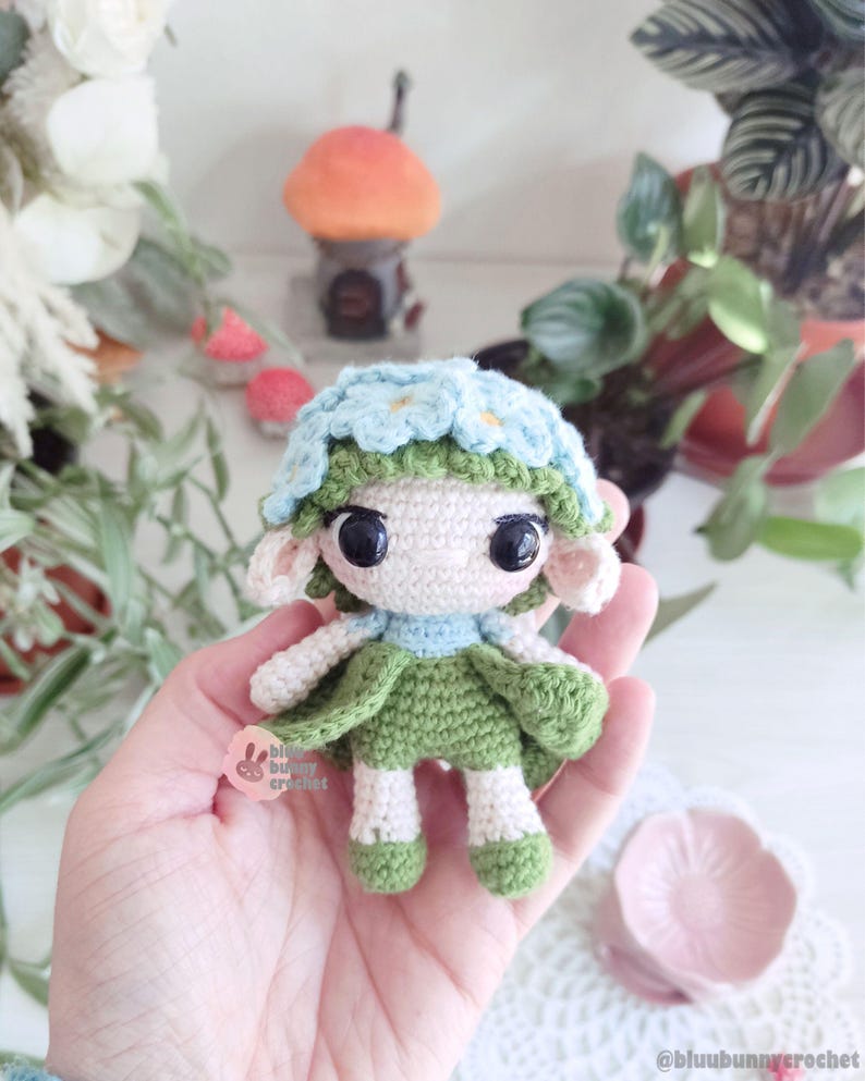 Puede incluir: Mu&ntilde;eca de ganchillo hecha a mano con una corona de flores y un vestido verde. La mu&ntilde;eca tiene ojos negros grandes, orejas peque&ntilde;as y un cuello azul claro. La mu&ntilde;eca se sostiene en una mano, con un plato rosa en forma de flor en el fondo.