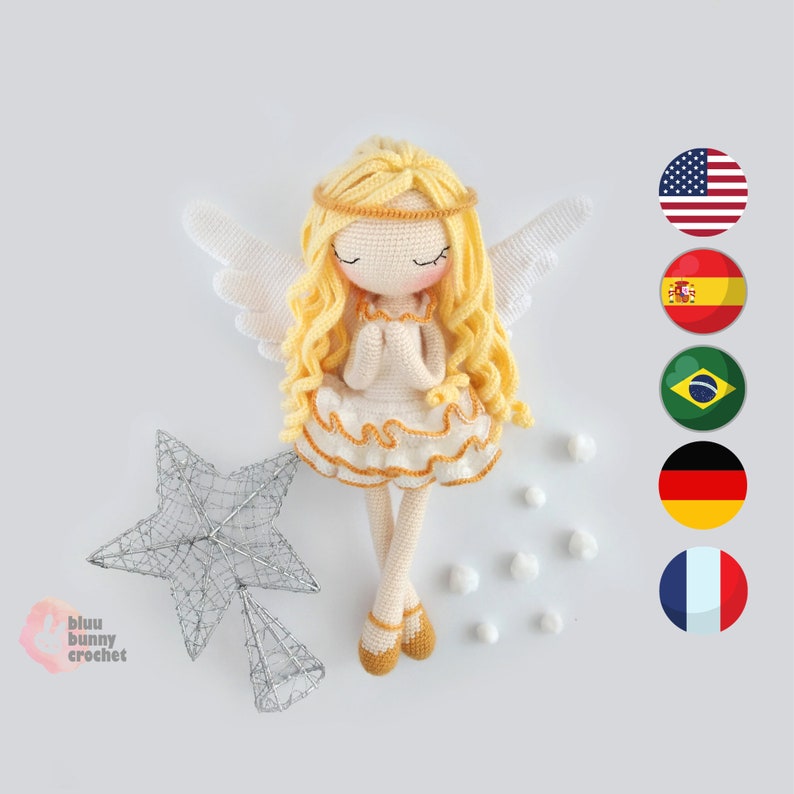 May include: A crocheted angel doll with blonde hair, white wings, and a white dress with a yellow trim. The doll is sitting on a white surface with a silver star and white pom-poms.