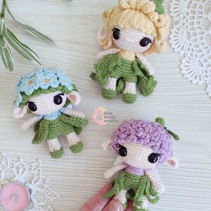 Puede incluir: Tres mu&ntilde;ecas de ganchillo hechas a mano con pelo en forma de flor y faldas verdes. Una mu&ntilde;eca tiene el pelo azul claro con detalles de flores azules, otra tiene el pelo rubio y la tercera tiene el pelo morado. Las mu&ntilde;ecas tienen ojos negros grandes.