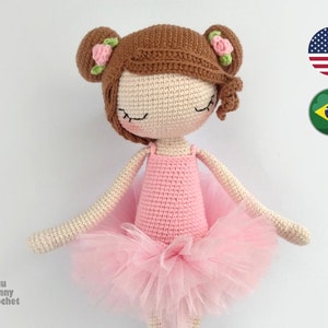 Może przedstawiać: Lalka balerina wykonana na szydełku z brązowymi włosami, zamkniętymi oczami i różową spódniczką. Lalka ma na sobie różową sukienkę wykonaną na szydełku i ma po jednej kwiecie z każdej strony głowy.