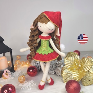 May include: A crocheted elf doll with brown curly hair, wearing a red dress with a green bodice, a red hat, and red shoes. The doll is standing on a wooden surface with Christmas decorations.