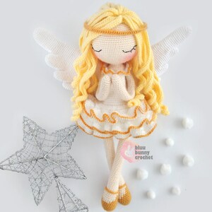 May include: A crocheted angel doll with long blonde hair, wearing a white dress with a yellow trim and a gold halo. The doll is sitting with its hands clasped in prayer. The doll is surrounded by white cotton balls and a silver star.