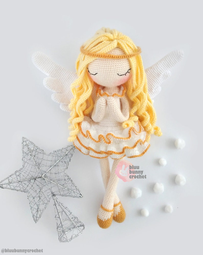 May include: A crocheted angel doll with long blonde hair, wearing a white dress with a yellow trim and a gold halo. The doll is sitting with its hands clasped in prayer. The doll is surrounded by white cotton balls and a silver star.