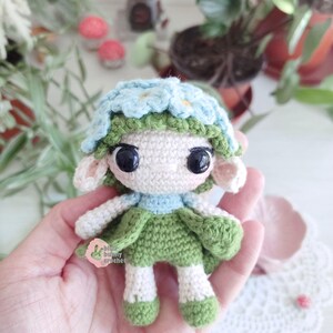 Puede incluir: Mu&ntilde;eca de ganchillo hecha a mano con una corona de flores y un vestido verde. La mu&ntilde;eca tiene ojos negros grandes, orejas peque&ntilde;as y un cuello azul claro. La mu&ntilde;eca mide aproximadamente 10 cm de altura.