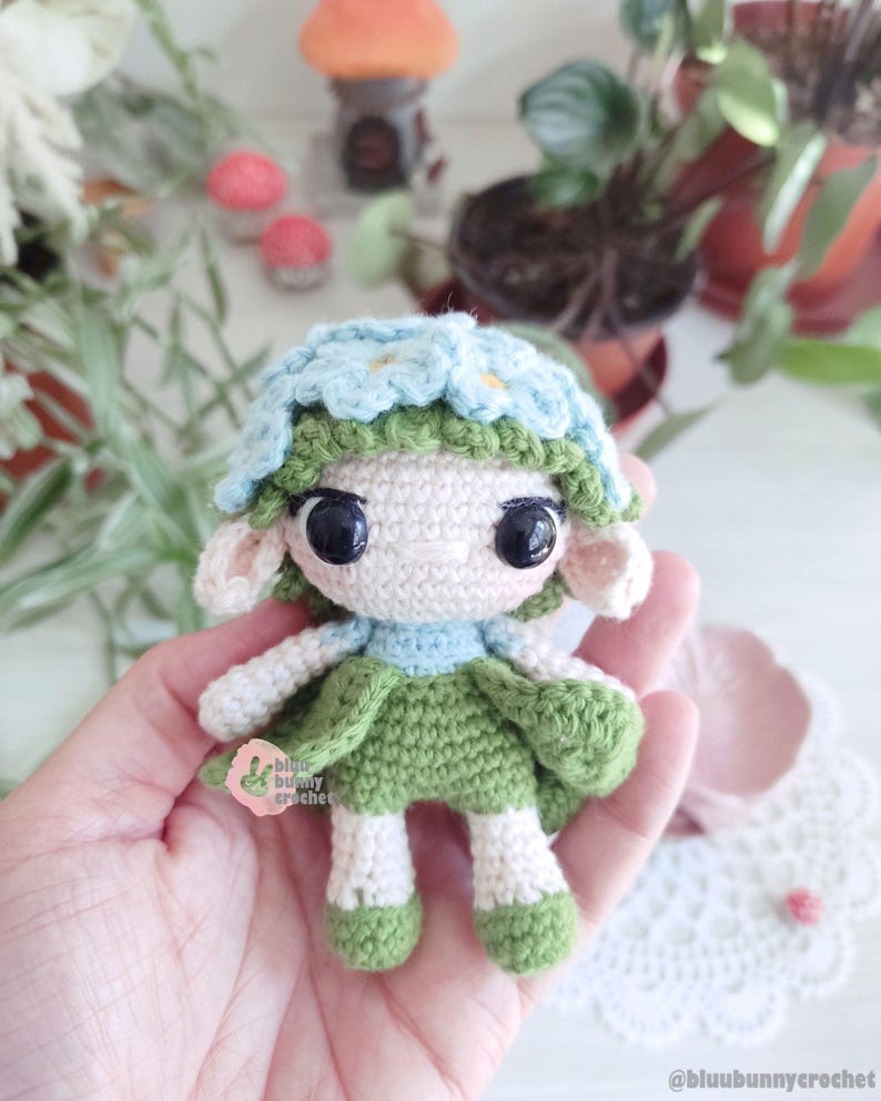 Puede incluir: Mu&ntilde;eca de ganchillo hecha a mano con una corona de flores y un vestido verde. La mu&ntilde;eca tiene ojos negros grandes, orejas peque&ntilde;as y un cuello azul claro. La mu&ntilde;eca mide aproximadamente 10 cm de altura.