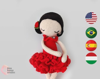 Puede incluir: Muñeca de crochet con un vestido rojo con volantes y una flor roja en el pelo. La muñeca tiene el pelo negro y los ojos cerrados. La muñeca está sentada sobre un fondo blanco.