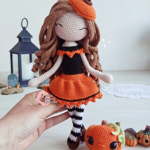 May include: Crocheted doll dressed in an orange and black pumpkin costume with a brown beret. The doll is holding a small crocheted pumpkin with a green stem and black eyes. The doll is standing on a white surface with other small pumpkins.