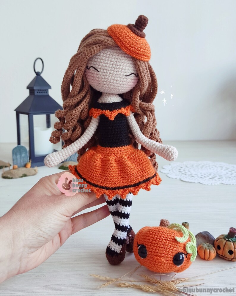 May include: Crocheted doll dressed in an orange and black pumpkin costume with a brown beret. The doll is holding a small crocheted pumpkin with a green stem and black eyes. The doll is standing on a white surface with other small pumpkins.