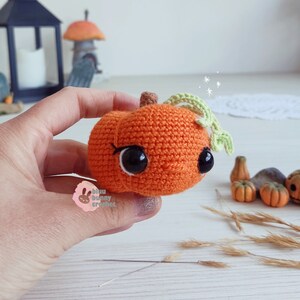 May include: A crocheted orange pumpkin with a green stem and leaves. The pumpkin has black eyes and a stitched smile. The pumpkin is being held in a hand.