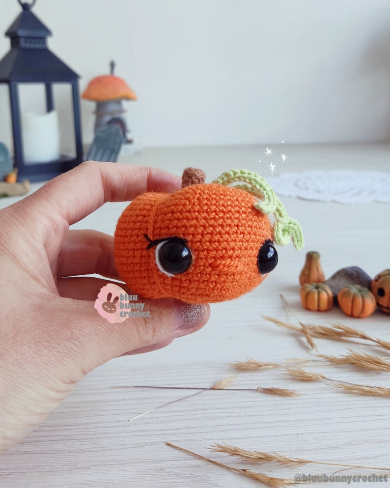 May include: A crocheted orange pumpkin with a green stem and leaves. The pumpkin has black eyes and a stitched smile. The pumpkin is being held in a hand.