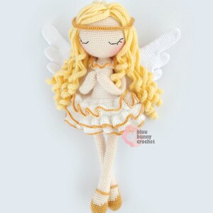 May include: A crocheted angel doll with long blonde hair, a white dress with gold trim, and white wings. The doll is wearing gold shoes and has its eyes closed.