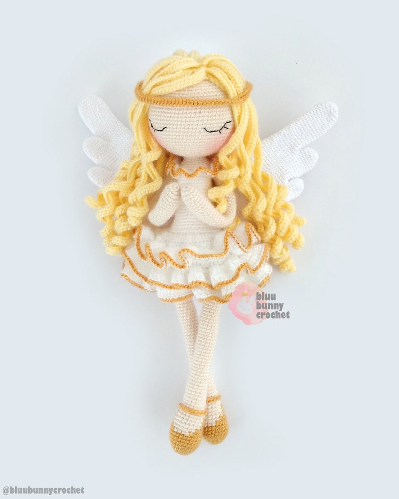 May include: A crocheted angel doll with long blonde hair, a white dress with gold trim, and white wings. The doll is wearing gold shoes and has its eyes closed.