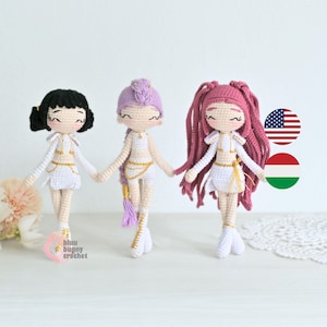 以下が含まれることがあります： 黒、ラベンダー、ピンクの髪の毛を持つ、手作りのかぎ針編み人形が3体。それぞれの人形は、金のアクセントが付いた白い衣装を着ており、お互いに手をつないでいます。人形は目を閉じて頬がバラ色です。人形は白い表面に立っています。