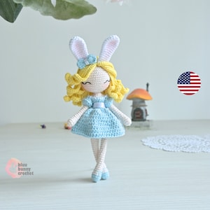 May include: A crocheted doll with blonde hair, a blue dress, and white bunny ears. The doll is standing on a white surface with a doily in the background.