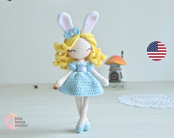 May include: A crocheted doll with blonde hair, a blue dress, and white bunny ears. The doll is standing on a white surface with a doily in the background.