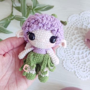 Puede incluir: Una mu&ntilde;eca de ganchillo hecha a mano con una cabeza lavanda, una cara blanca y ojos negros grandes. La mu&ntilde;eca tiene brazos blancos, una parte superior lavanda y pantalones verdes con flecos. La mu&ntilde;eca sostiene una peque&ntilde;a etiqueta rosa con el texto "blue bub crochet".