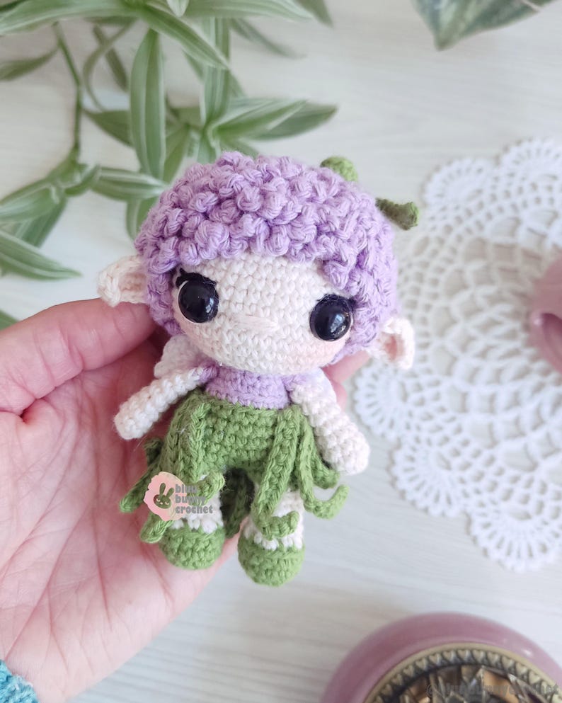 Puede incluir: Una mu&ntilde;eca de ganchillo hecha a mano con una cabeza lavanda, una cara blanca y ojos negros grandes. La mu&ntilde;eca tiene brazos blancos, una parte superior lavanda y pantalones verdes con flecos. La mu&ntilde;eca sostiene una peque&ntilde;a etiqueta rosa con el texto "blue bub crochet".
