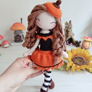 May include: Crocheted doll with brown hair, wearing an orange and black dress, a pumpkin hat, and striped stockings. The doll is standing on a wooden surface with a sunflower in the background.