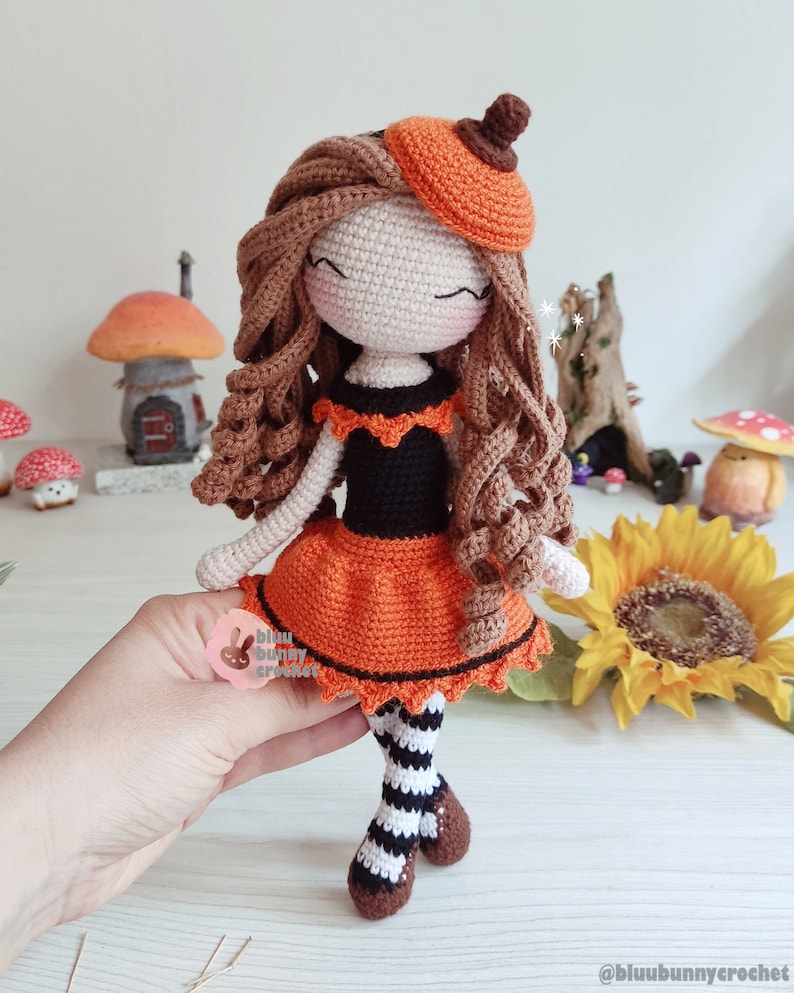 May include: Crocheted doll with brown hair, wearing an orange and black dress, a pumpkin hat, and striped stockings. The doll is standing on a wooden surface with a sunflower in the background.