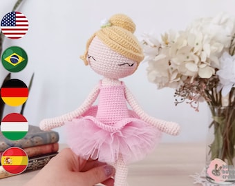 May include: A crocheted ballerina doll with blonde hair, a pink dress, and a tulle skirt. The doll is standing on a table with a bouquet of white flowers in the background.
