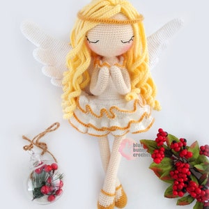 May include: A crocheted angel doll with long blonde hair, white wings, and a white dress with gold trim. The doll is sitting with its hands clasped in prayer. The doll is on a white background with a small glass ornament and a sprig of red berries.