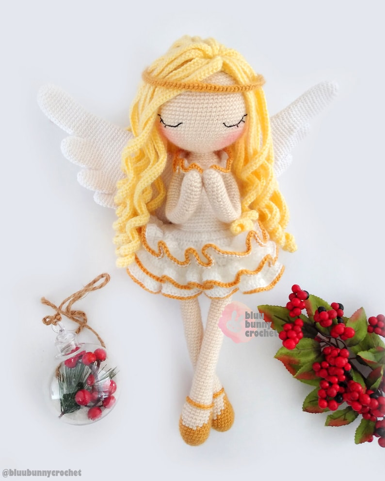 May include: A crocheted angel doll with long blonde hair, white wings, and a white dress with gold trim. The doll is sitting with its hands clasped in prayer. The doll is on a white background with a small glass ornament and a sprig of red berries.